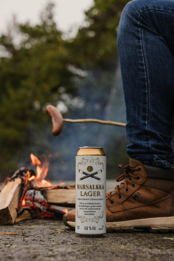 Marsalkka lager tölkki nuotion edessä, taustalla makkara grillitikussa